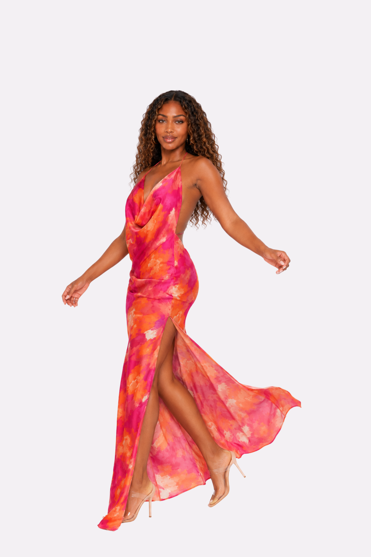 A woman modeling a vibrant, one-shoulder maxi dress with a pink, orange, and yellow tie-dye pattern, set against a solid light gray background.