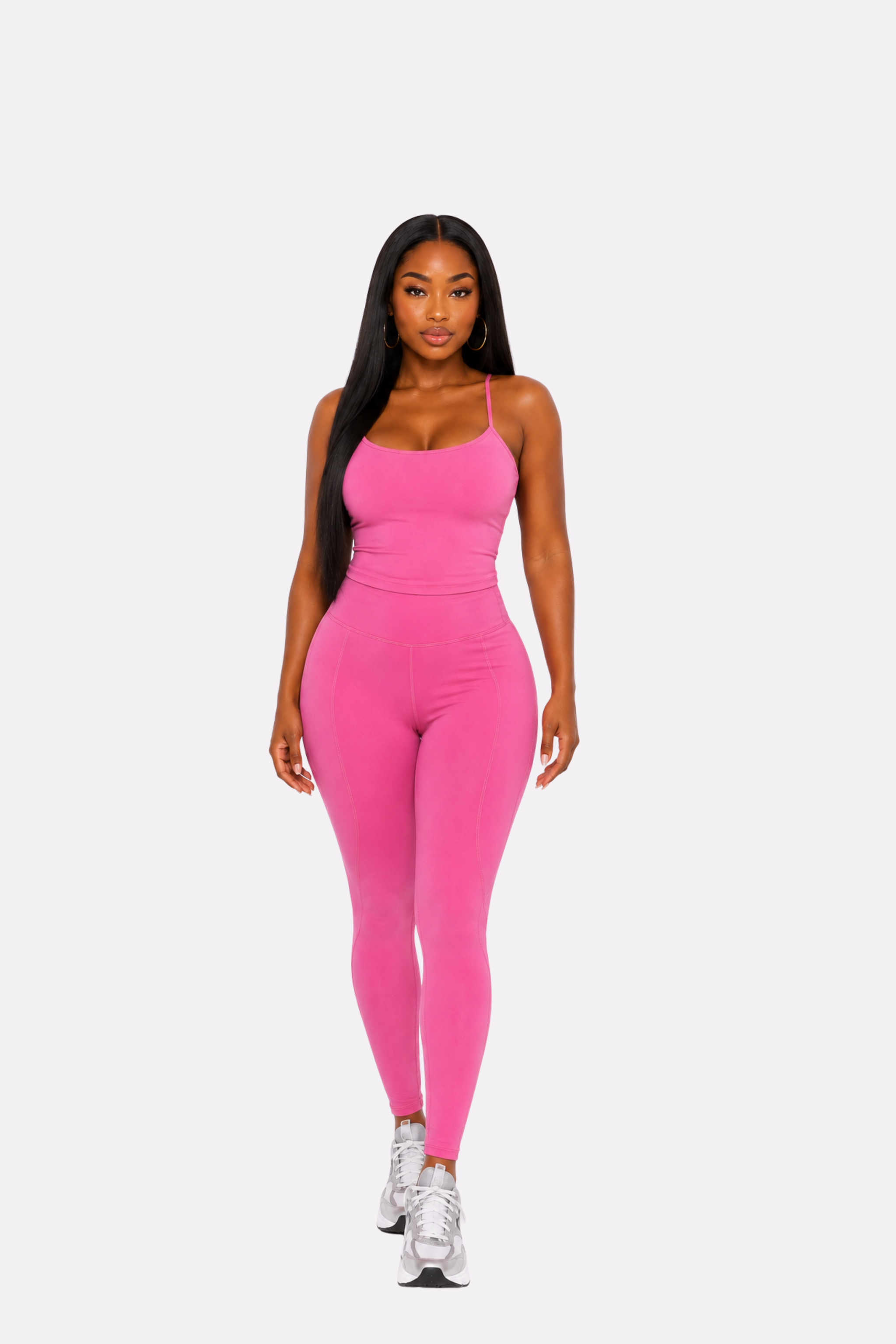 A woman modeling a bright pink two-piece activewear set consisting of a spaghetti-strap tank top and matching high-waisted leggings, paired with white sneakers, against a solid light gray background.