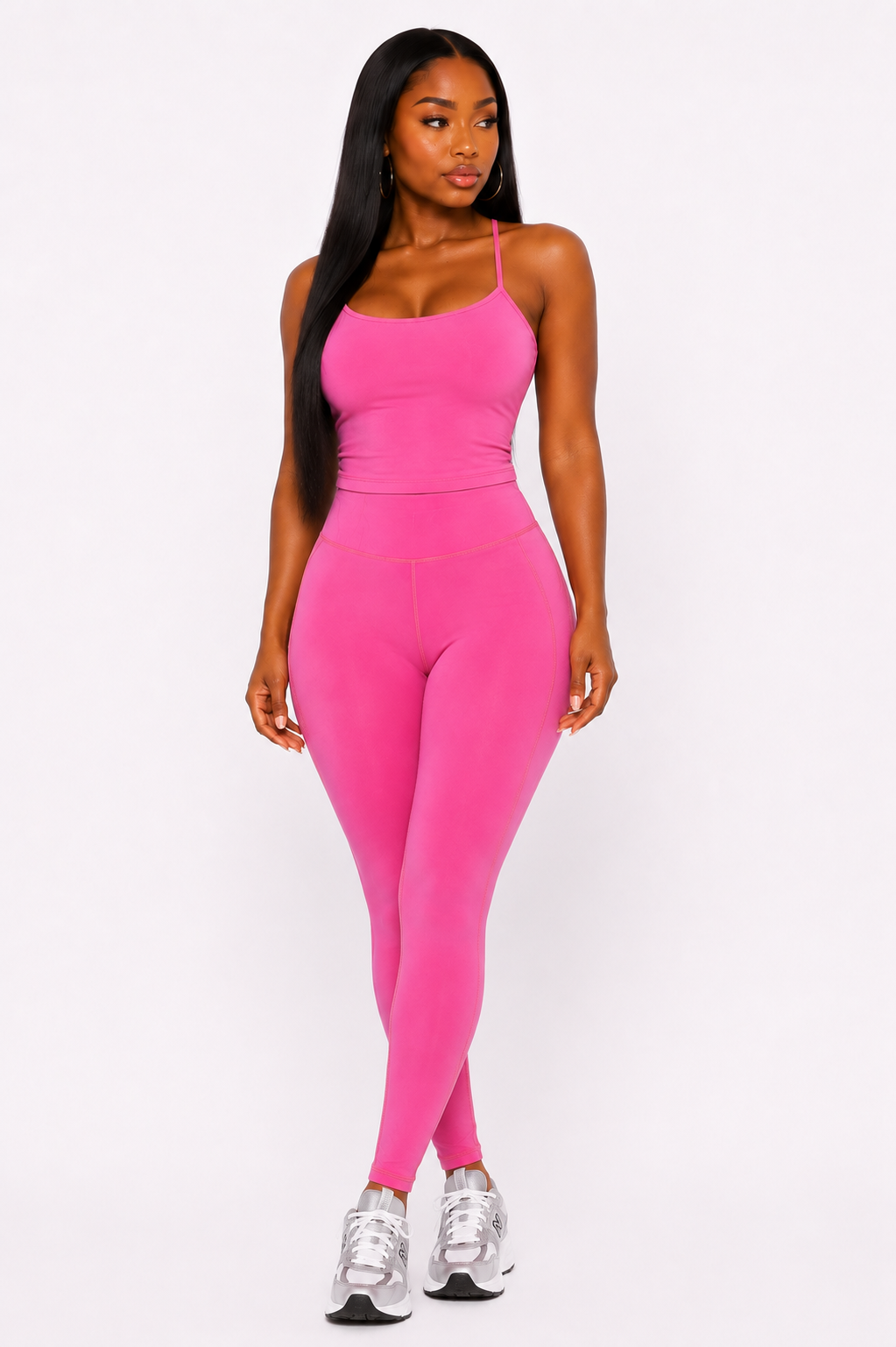 A woman modeling a bright pink two-piece activewear set consisting of a spaghetti-strap tank top and matching high-waisted leggings, paired with silver sneakers, against a plain white studio background.