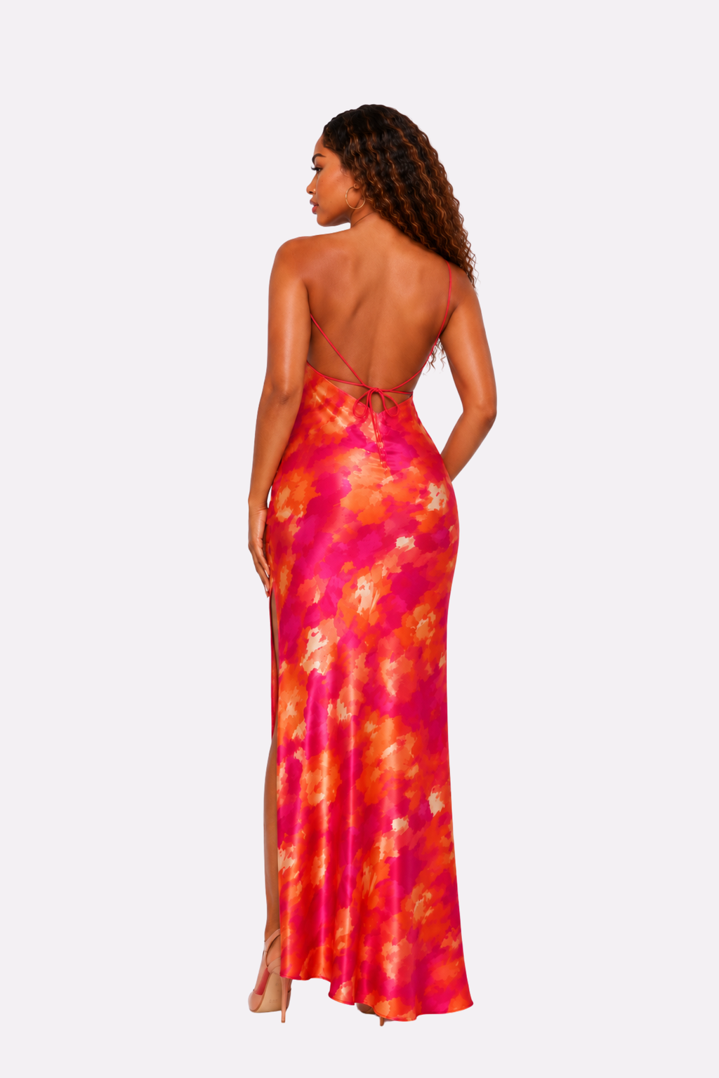 A woman shown from the back wearing a sleeveless, open-back maxi dress with a vibrant pink, orange, and yellow floral tie-dye pattern, set against a solid light gray background.