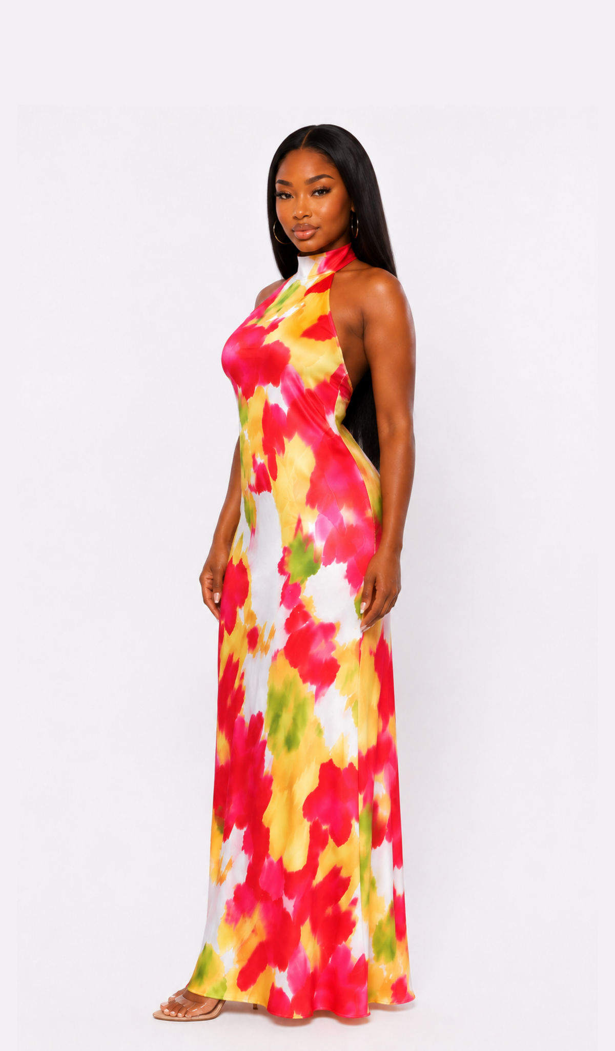 A woman modeling a sleeveless halter-neck maxi dress with a vibrant watercolor-style abstract print in red, yellow, green, and white, set against a plain light gray studio background.