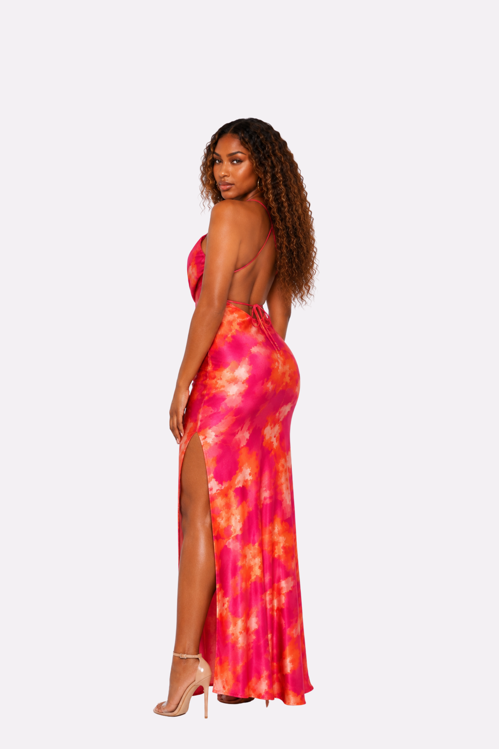 A woman modeling a vibrant pink and orange tie-dye maxi dress with a backless open back and high leg slit, shown from a three-quarters back view against a light gray background.