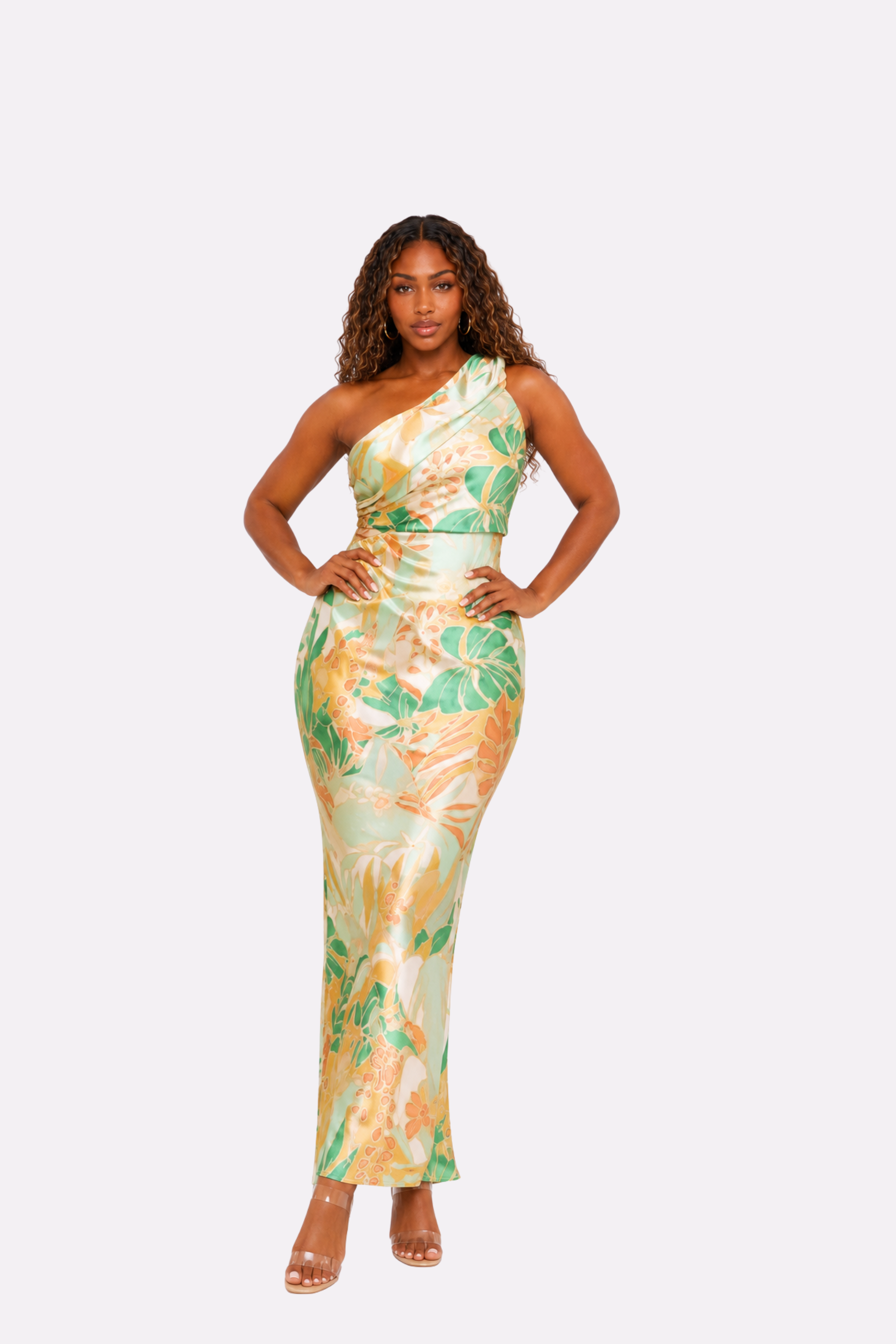 A woman modeling a one-shoulder, floor-length satin dress with a tropical leaf print in green, yellow, and beige, set against a solid light gray background.
