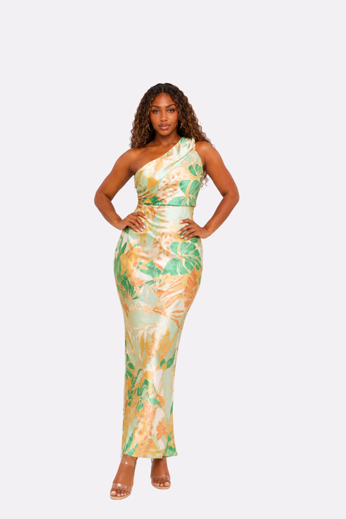 A woman modeling a one-shoulder, floor-length satin dress with a tropical leaf print in green, yellow, and beige, set against a solid light gray background.