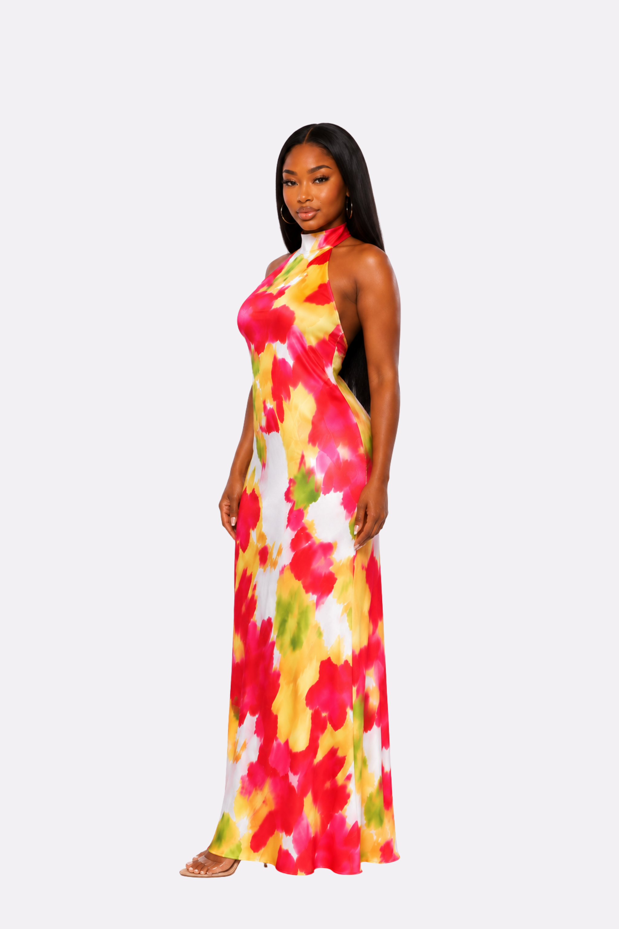 A woman modeling a sleeveless halter-neck maxi dress with a vibrant abstract watercolor print in red, yellow, green, and white, set against a solid light gray background.