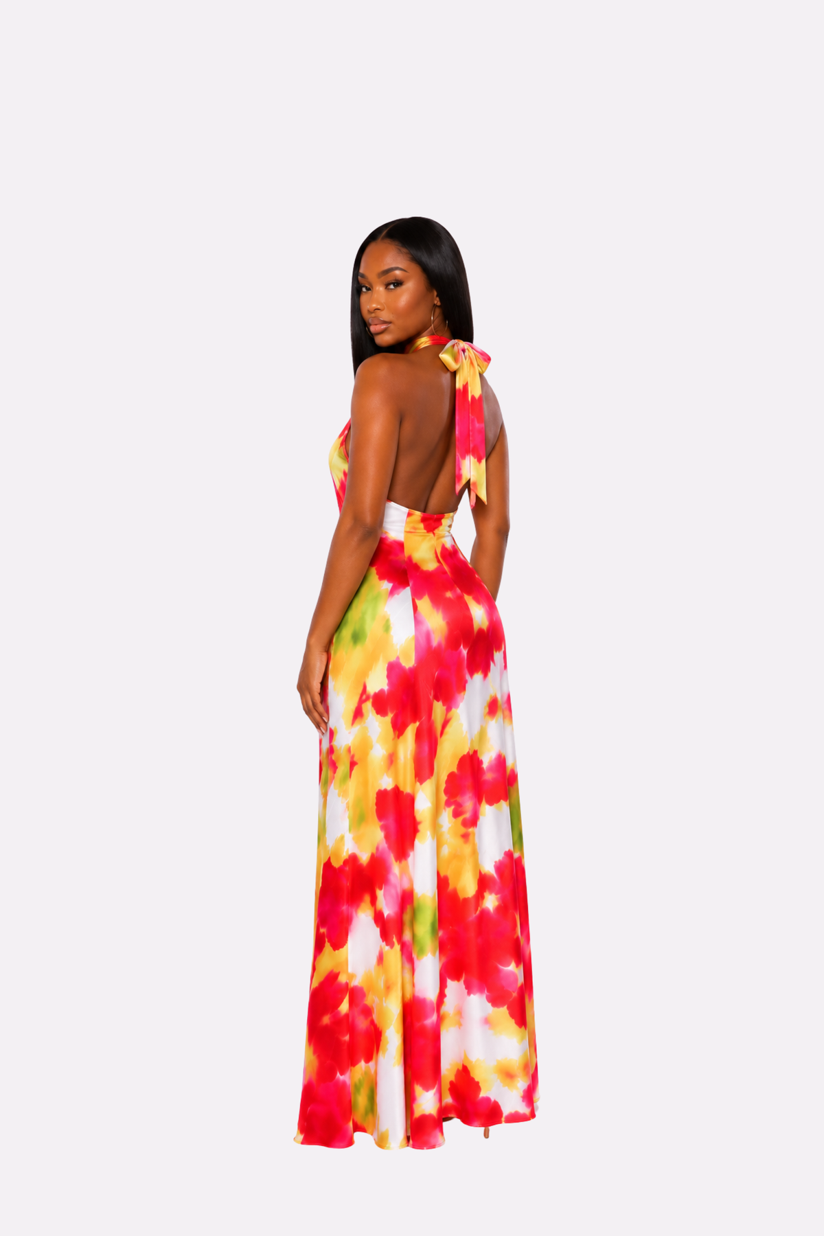 A woman modeling a long halter maxi dress from the back, featuring a vibrant red, yellow, and green tie-dye floral pattern on a white base, set against a plain light gray background.